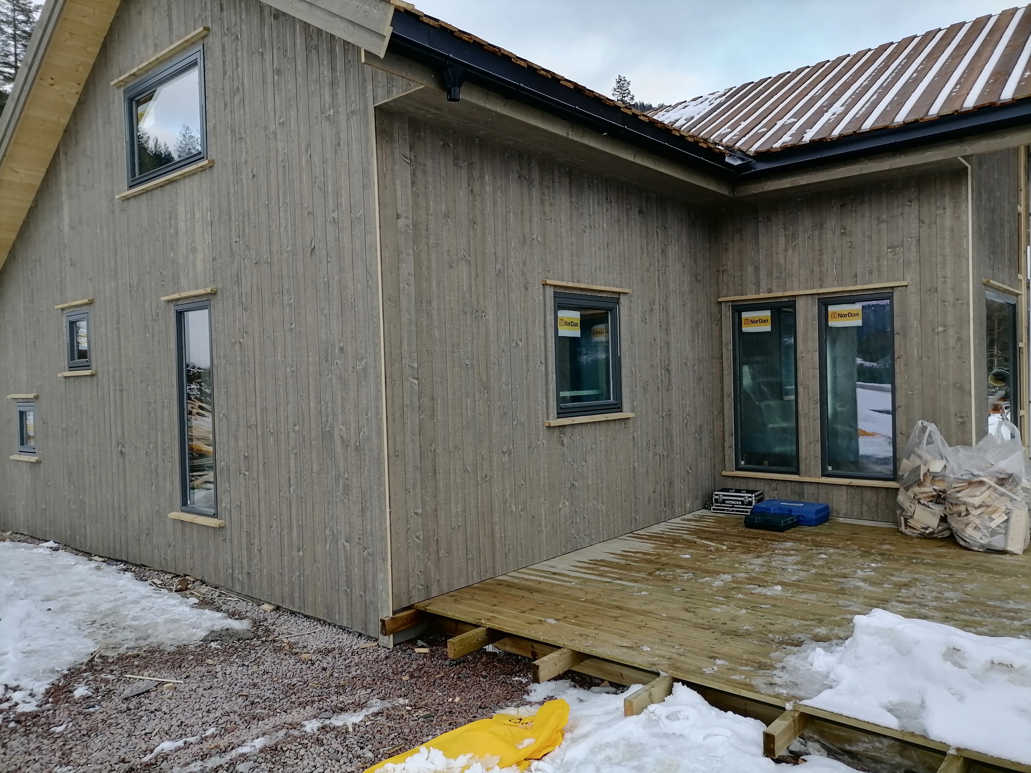 Nowoczesny dom z szarą drewnianą elewacją, czarnymi rynnami i częściowo ośnieżonym dachem z drewnianych desek, widoczne okna z naklejkami producenta oraz drewniany taras w trakcie budowy.