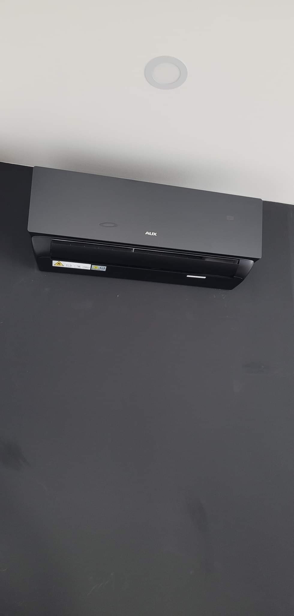 Czarny klimatyzator AUX zamontowany na ciemnoszarej ścianie w pomieszczeniu z białym sufitem i okrągłym oświetleniem punktowym.