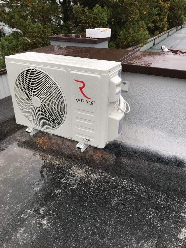 Biała jednostka zewnętrzna klimatyzatora Rotenso zamontowana na płaskim, mokrym dachu z widocznymi śladami brudu i pyłu, z drzewami w tle.