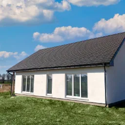 Nowoczesny, parterowy budynek z białym tynkiem, ciemną dachówką i dużymi oknami na tle błękitnego nieba z chmurami.