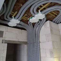 Szare, karbowane rury wentylacyjne rozprowadzone pod sufitem w surowym stanie, z widocznymi elementami mocującymi i izolacją, w budynku z białego bloczka.