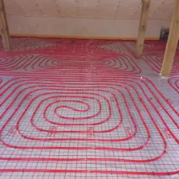 Czerwone rurki ogrzewania podłogowego ułożone spiralnie na siatce, widoczne drewniane elementy konstrukcji dachu na poddaszu.