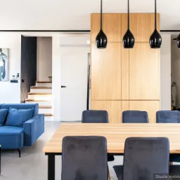 Jasne wnętrze z drewnianym stołem, szarymi krzesłami, niebieską sofą i wiszącymi czarnymi lampami; w tle zabudowa z jasnego drewna i schody.