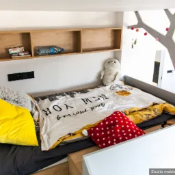 Pokój dziecięcy z antresolą: łóżko na podeście z drewnianymi półkami na książki i dekoracjami, w tym lampki w kształcie drzewa i pluszowe zabawki.