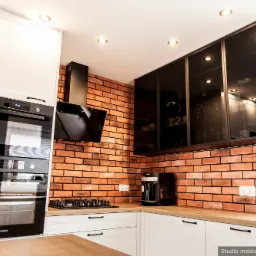 Nowoczesna kuchnia w Gliwicach z białymi frontami i ceglaną ścianą, wyposażona w piekarnik, płytę gazową, okap i przeszklone szafki wiszące.