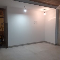 Jacek M - Wnętrze w trakcie remontu: białe ściany po malowaniu, surowy betonowy strop z widocznymi instalacjami oświetleniowymi i szarym obramowaniem, szara listwa przypodłogowa, oraz szara podłoga. W tle...