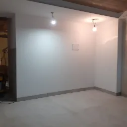 Wnętrze w trakcie remontu: białe ściany po malowaniu, surowy betonowy strop z widocznymi instalacjami oświetleniowymi i szarym obramowaniem, szara listwa przypodłogowa, oraz szara podłoga. W tle...