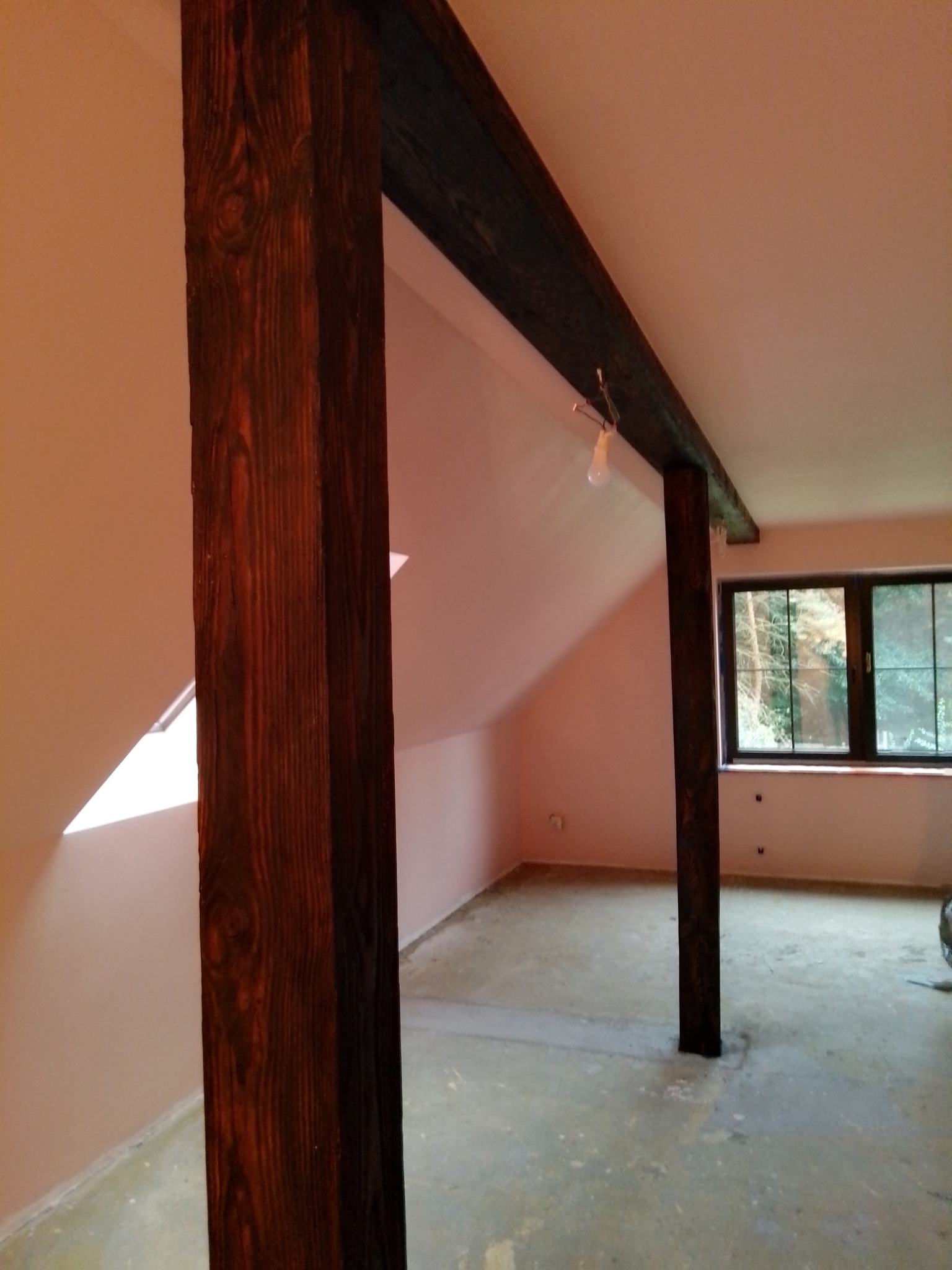 Wnętrze poddasza z drewnianą konstrukcją nośną, widoczne słupy i belka w ciemnym brązie, surowy beton na podłodze, okno z widokiem na zieleń.