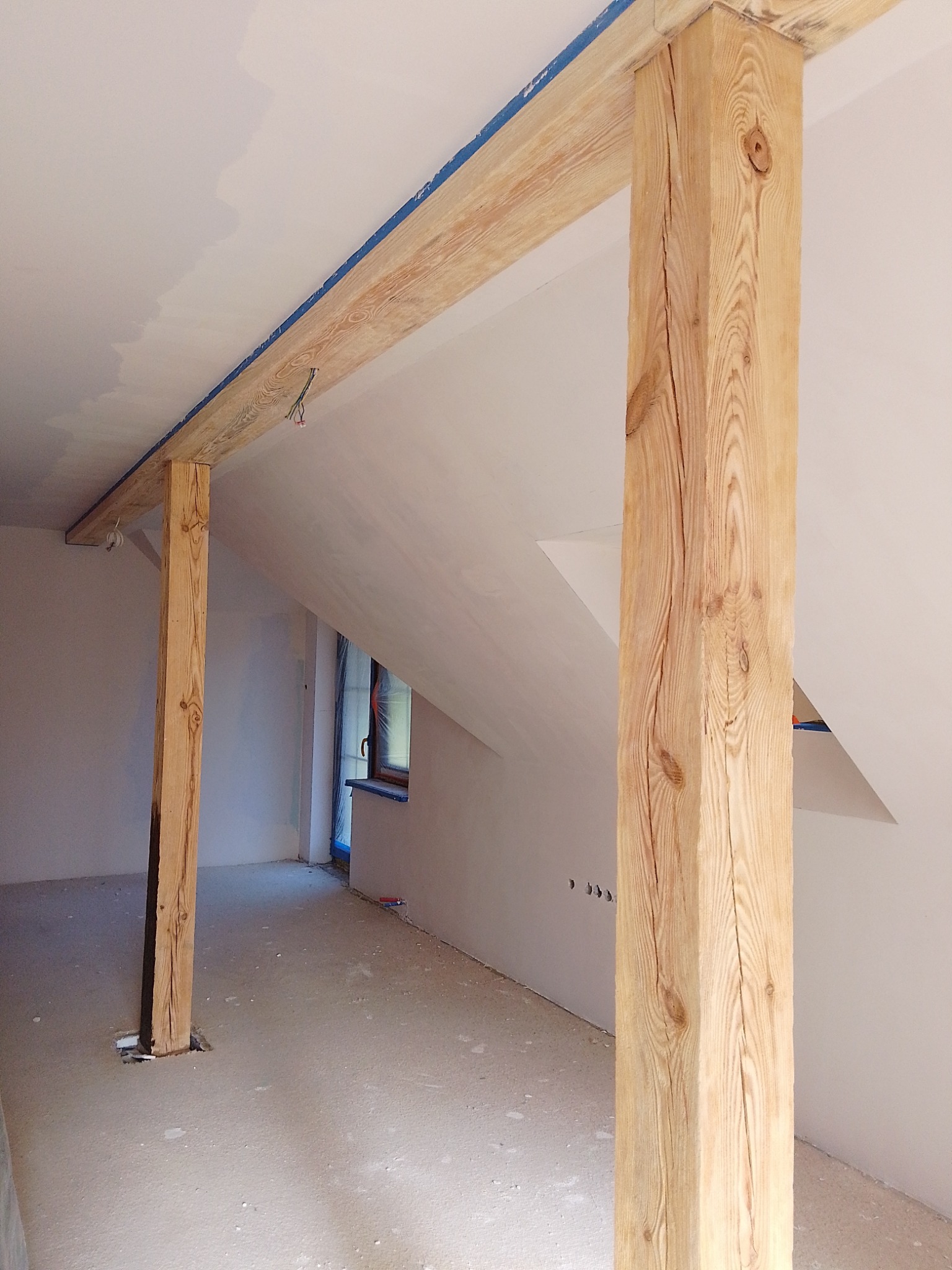 Poddasze w trakcie remontu: drewniane belki konstrukcyjne z widocznym uszczelnieniem taśmą malarską, ściany po szpachlowaniu, przygotowane do malowania.