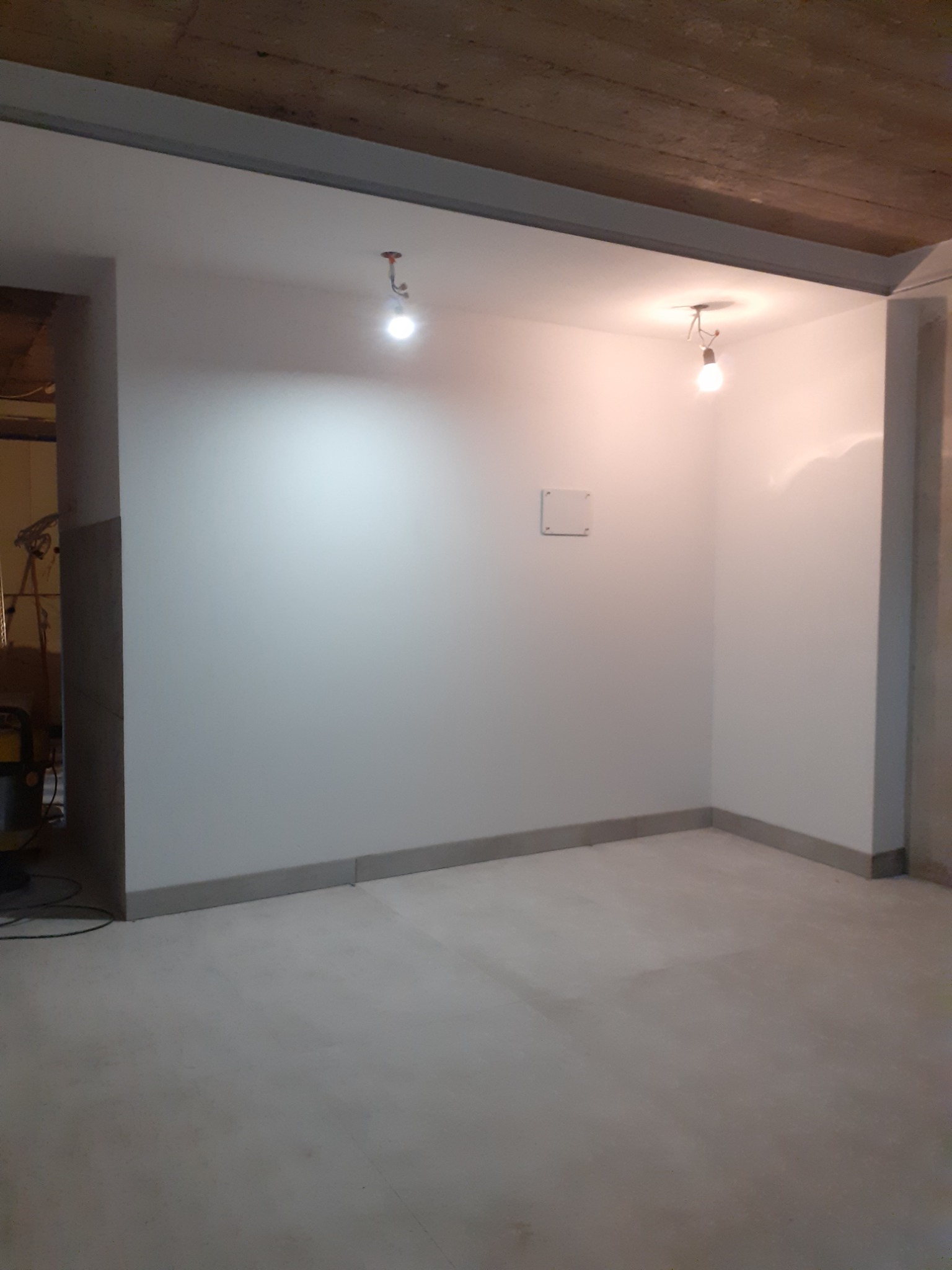 Wnętrze w trakcie remontu: białe ściany po malowaniu, surowy betonowy strop z widocznymi instalacjami oświetleniowymi i szarym obramowaniem, szara listwa przypodłogowa, oraz szara podłoga. W tle...