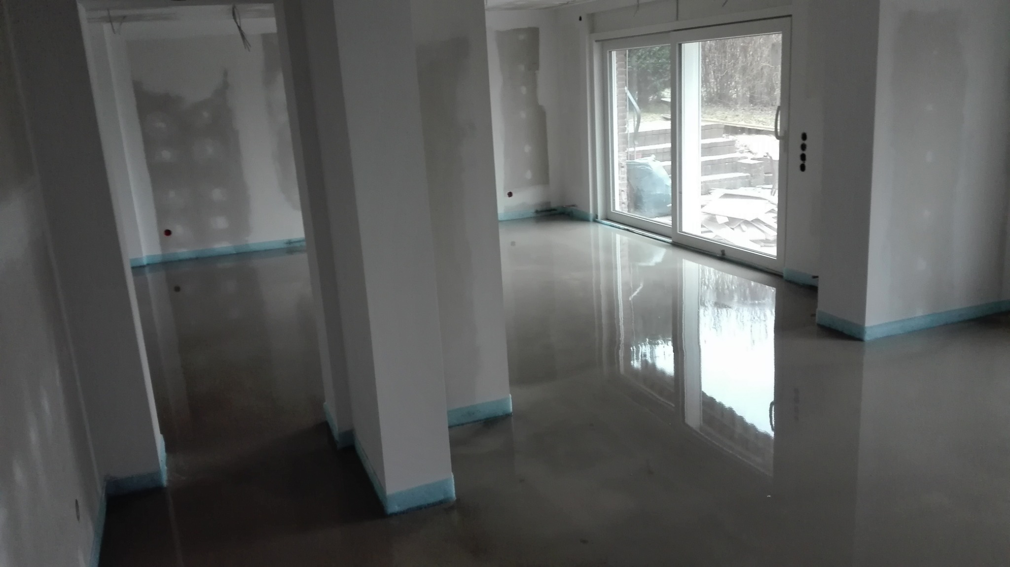 Wnętrze w trakcie remontu z wylaną posadzką samopoziomującą, zabezpieczone ściany i słupy, okno przesuwne z widokiem na ogród.