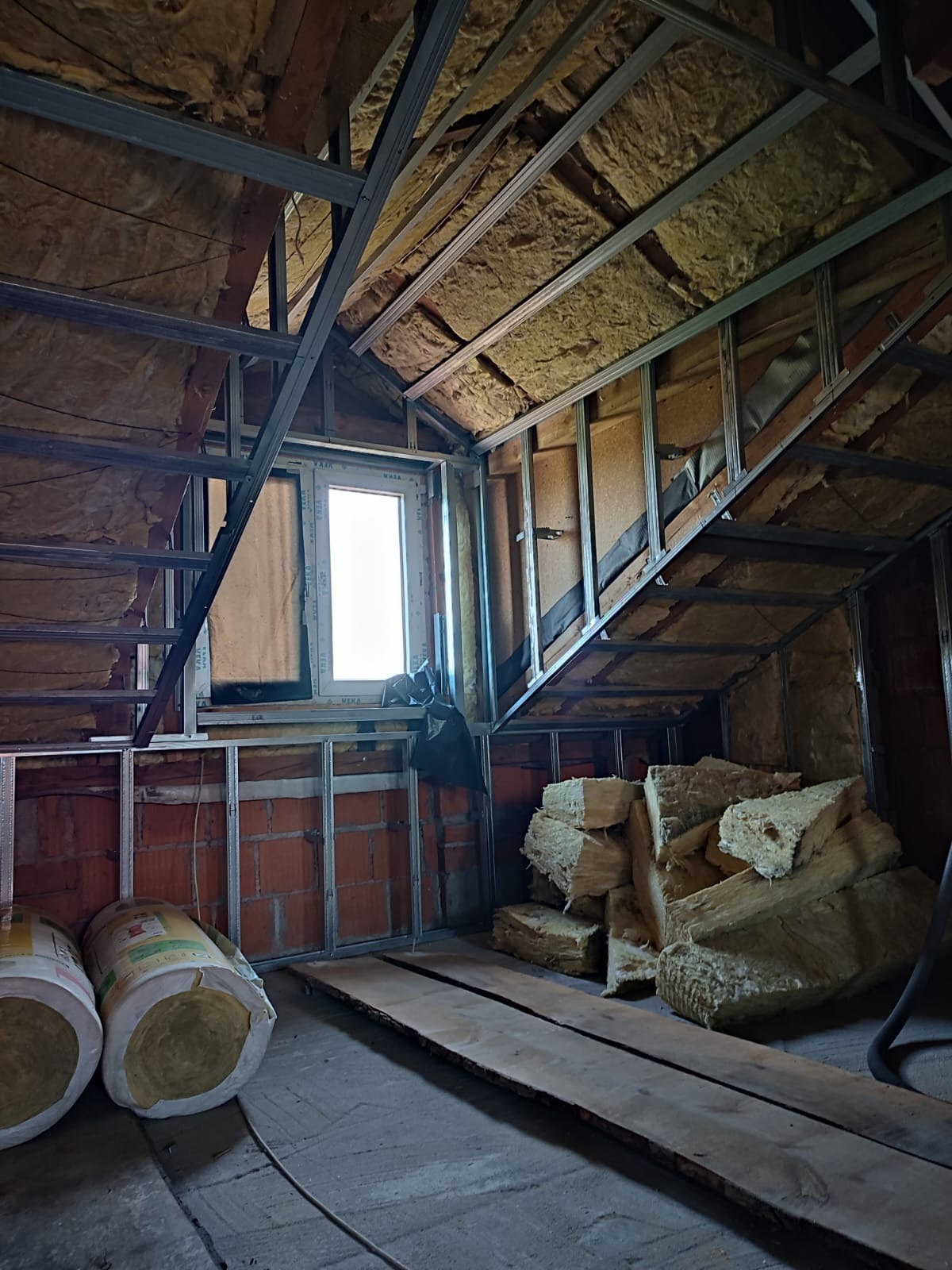 Poddasze w trakcie adaptacji: widoczne ocieplenie wełną mineralną, metalowy stelaż konstrukcyjny, ceglana ściana i skosy dachu z oknem.