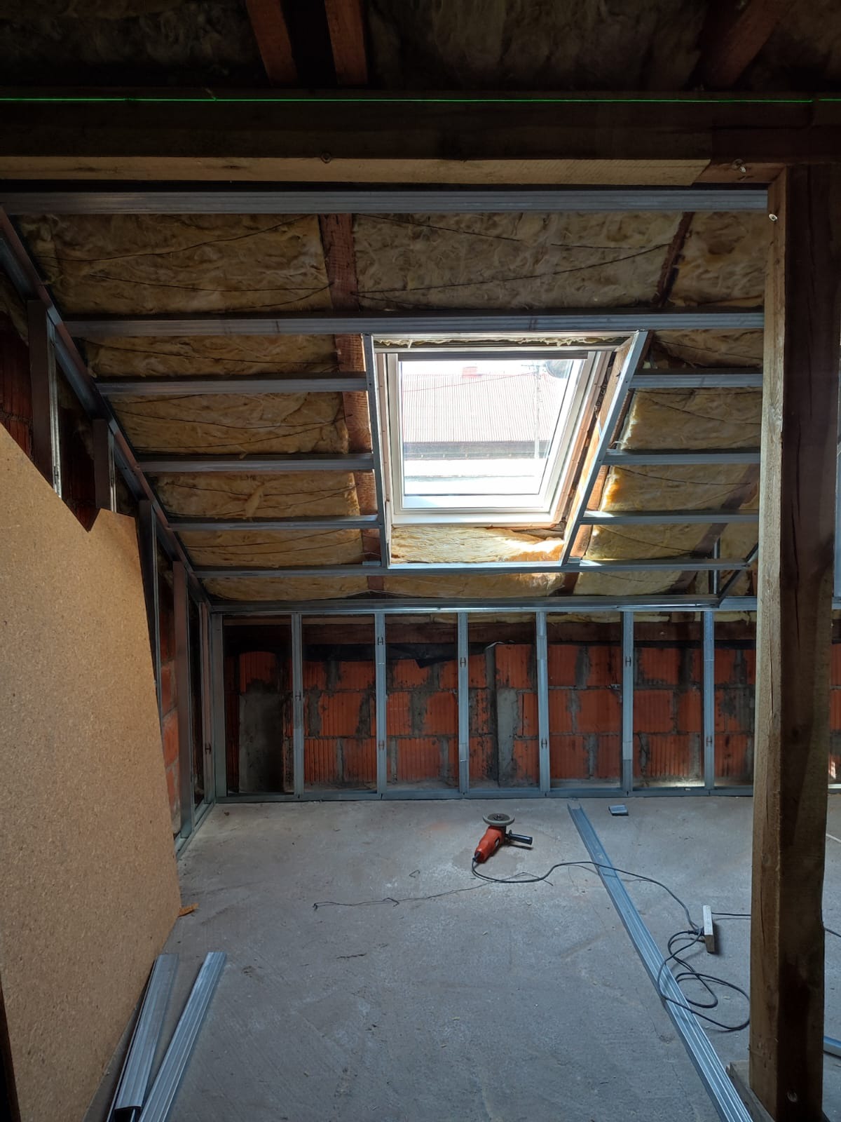 Stelaż z profili metalowych zamontowany na poddaszu z oknem dachowym, izolacja z wełny mineralnej widoczna w przestrzeni między krokwiami, ceglany mur w tle, szlifierka kątowa na podłodze.