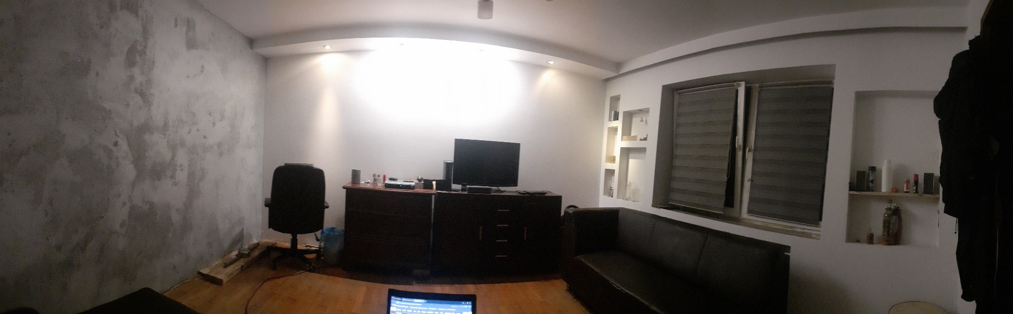 Pokój w trakcie remontu, z widoczną ścianą po wstępnym przygotowaniu pod malowanie lub wykończenie, meblami i sprzętem RTV w tle.