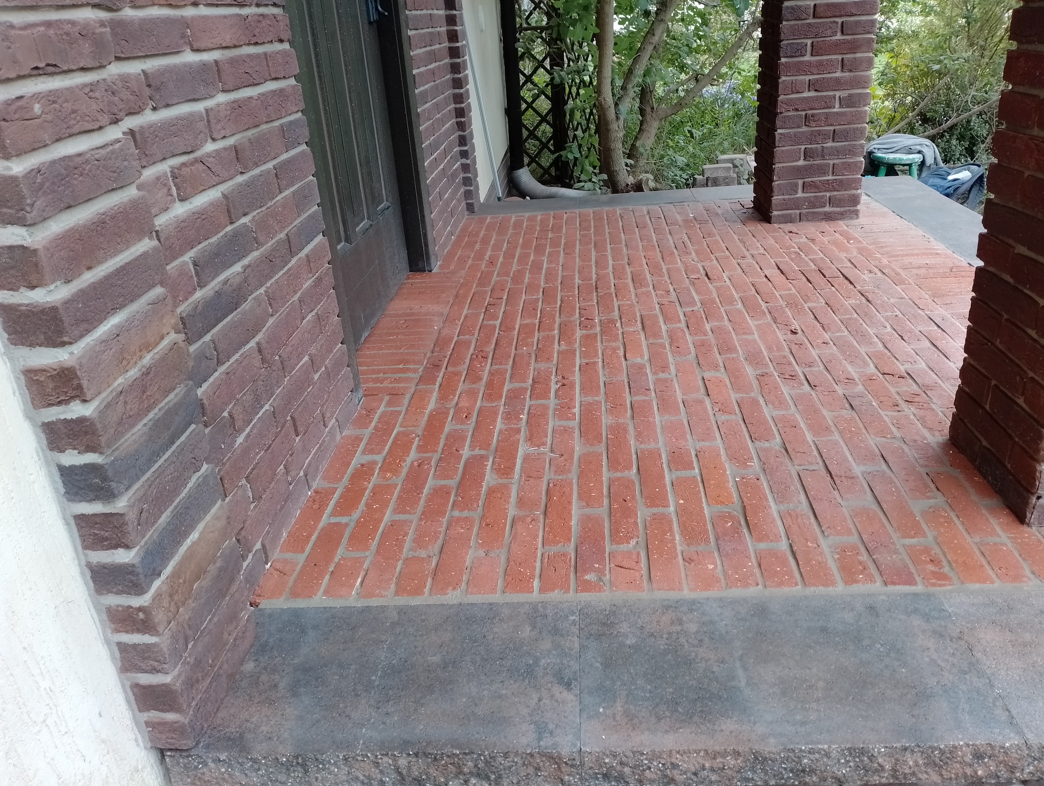 Ceglana weranda z ciemnymi fugami prowadząca do drzwi wejściowych, z kolumną z cegły po prawej stronie i widokiem na zieleń w tle.