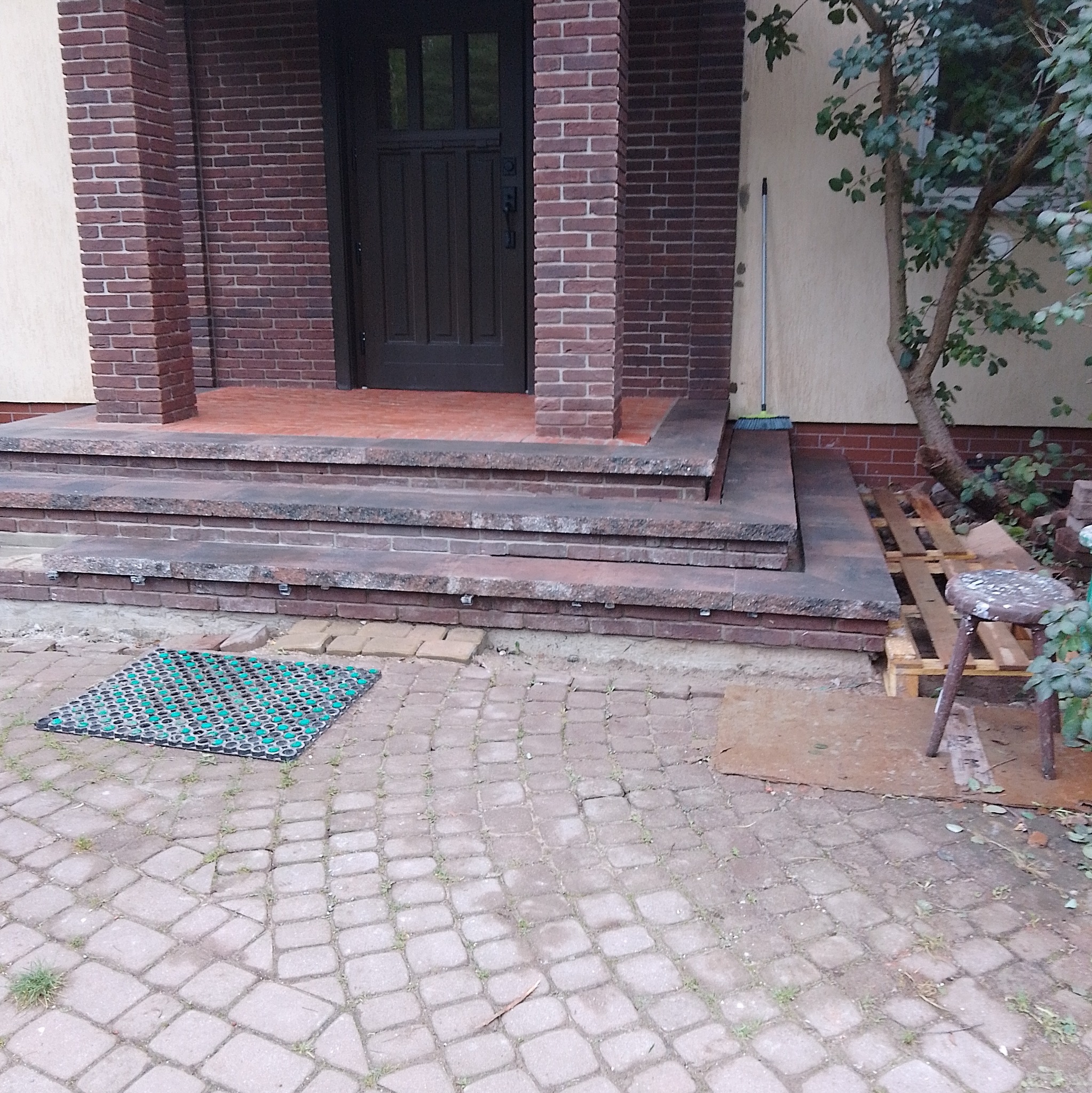 Wejście do domu z ciemnymi drzwiami, schodami wyłożonymi cegłą i kamienną kostką brukową na podwórku, z widocznym dywanikiem i małym stołkiem na palecie.