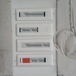 Szymon Wardin - Biała skrzynka rozdzielcza z zamontowanymi bezpiecznikami i przewodami elektrycznymi na surowej ścianie z betonu komórkowego.