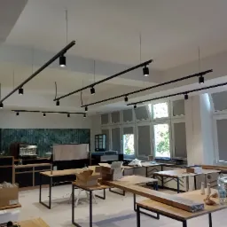 Wnętrze lokalu w trakcie wykańczania z zamontowanym systemem szynoprzewodów z reflektorami punktowymi, widoczne okna z roletami i meble w surowym stanie.