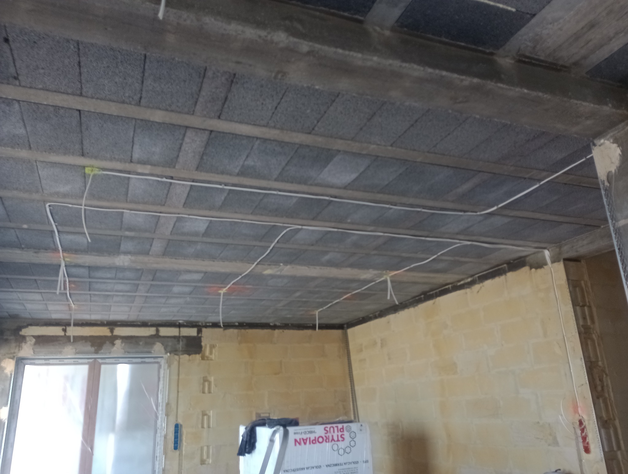 Białe kable elektryczne prowadzone pod betonowym stropem w surowym stanie, widoczne oznaczenia na kablach i suficie, ściany w trakcie wykańczania.