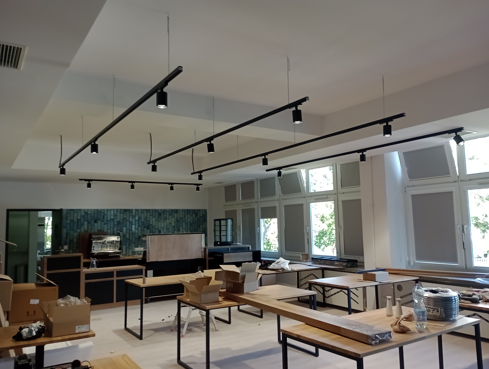 Wnętrze lokalu w trakcie wykańczania z zamontowanym systemem szynoprzewodów z reflektorami punktowymi, widoczne okna z roletami i meble w surowym stanie.