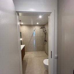 PHU POOLKRING Agnieszka Zenker - Nowoczesna łazienka z kabiną prysznicową typu walk-in, umywalką z szafką i toaletą. Beżowe płytki, czarna armatura, oświetlenie punktowe. Widok z korytarza.