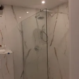 Nowoczesna kabina prysznicowa z deszczownicą i baterią podtynkową, wykończona płytkami imitującymi marmur z delikatnymi złotymi żyłkami, widoczny fragment toalety podwieszanej z niebieskimi...