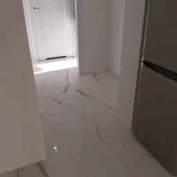 Nowe, jasne płytki podłogowe imitujące marmur z delikatnymi brązowymi żyłkami, widoczne w korytarzu z białą stolarką drzwiową i fragmentem szarej lodówki.