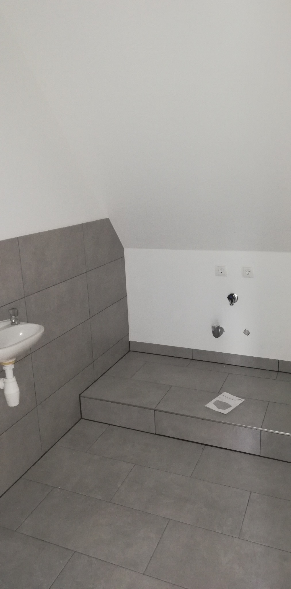Łazienka w trakcie wykańczania: szare płytki na ścianach i podłodze, zamontowana umywalka, wystające rury, widoczne gniazdka elektryczne, na podłodze leży kartka.