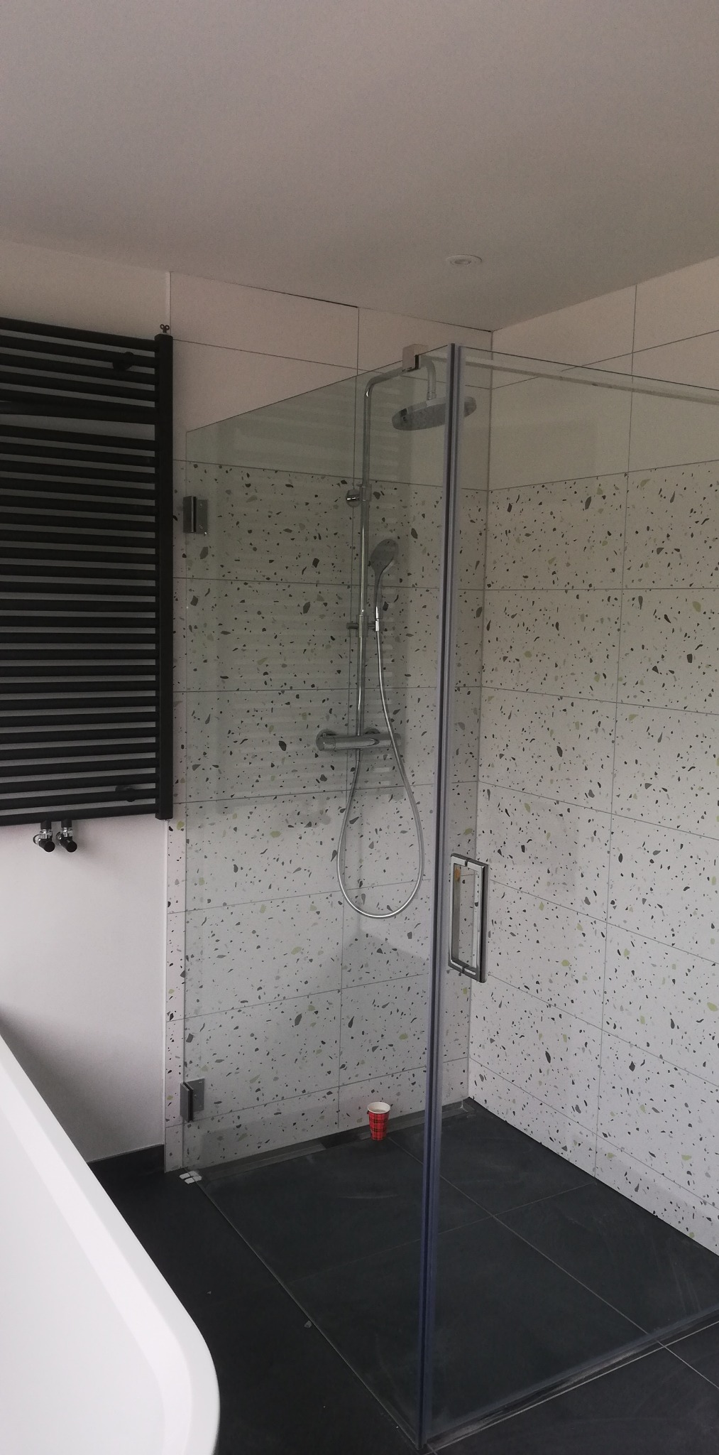 Nowoczesna kabina prysznicowa z transparentnego szkła z chromowanymi okuciami, jasne płytki z drobnymi, kolorowymi wzorami na ścianach, czarna podłoga, grzejnik drabinkowy w kolorze czarnym...