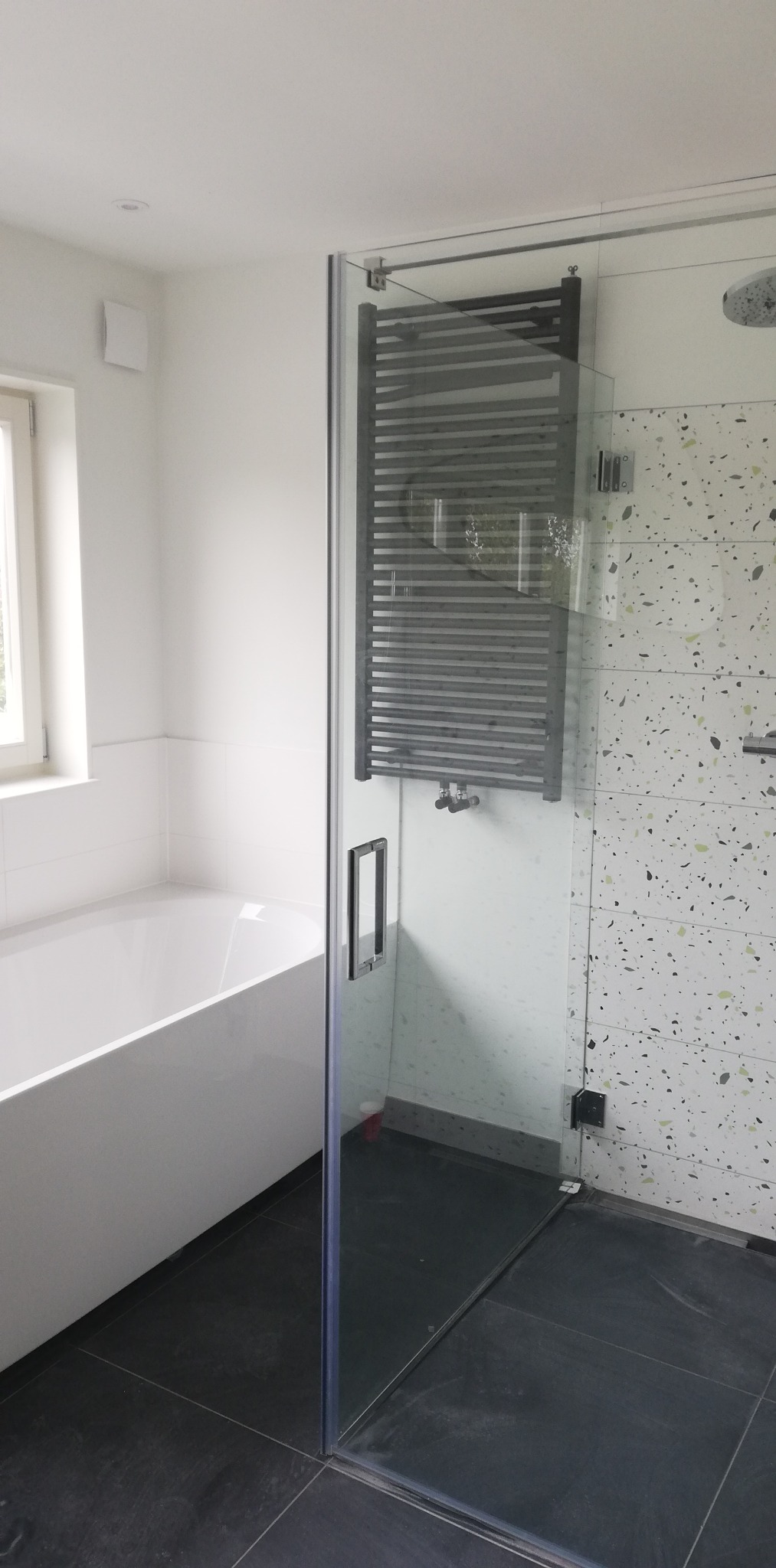 Nowoczesna łazienka z kabiną prysznicową ze szklanymi drzwiami, grzejnikiem drabinkowym na ścianie z mozaiką lastryko oraz czarną podłogą z płytek.