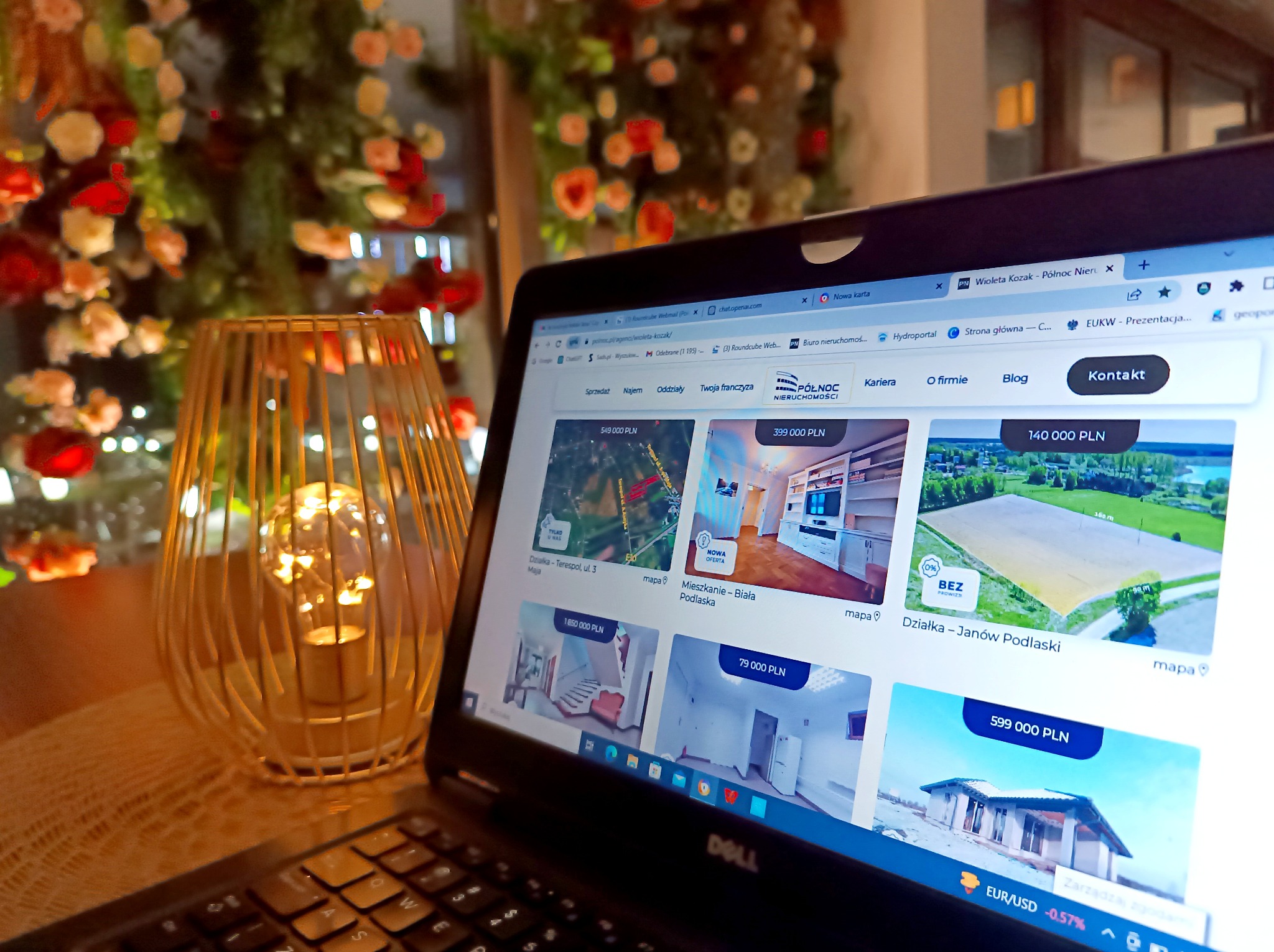 Laptop wyświetlający stronę internetową agencji nieruchomości z ofertami sprzedaży, obok dekoracyjna lampa z żarówką, w tle kwiatowa dekoracja.