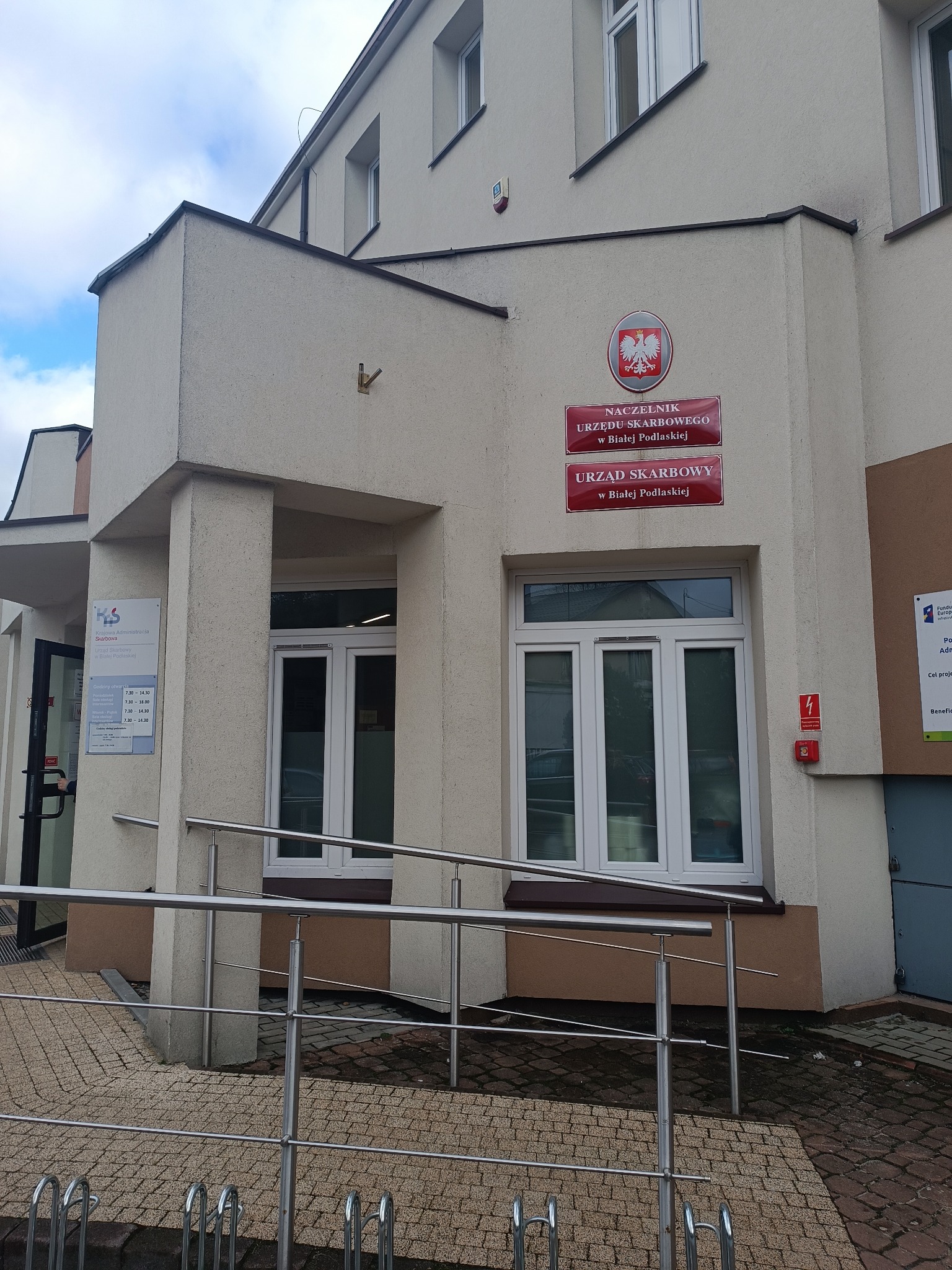 Budynek Urzędu Skarbowego w Białej Podlaskiej z widocznym wejściem dla osób niepełnosprawnych i tablicą informacyjną z godzinami otwarcia.