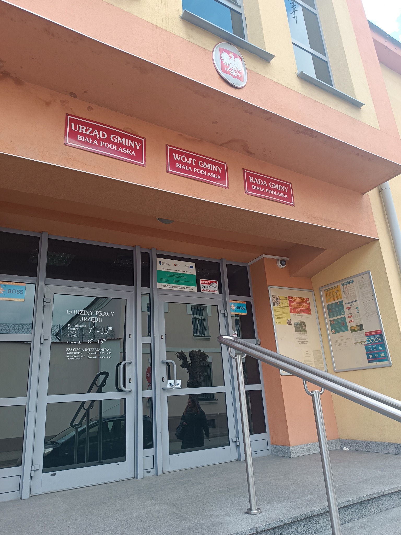 Wejście do Urzędu Gminy Biała Podlaska z widocznymi tablicami informacyjnymi, godzinami pracy urzędu i poręczami dla niepełnosprawnych.