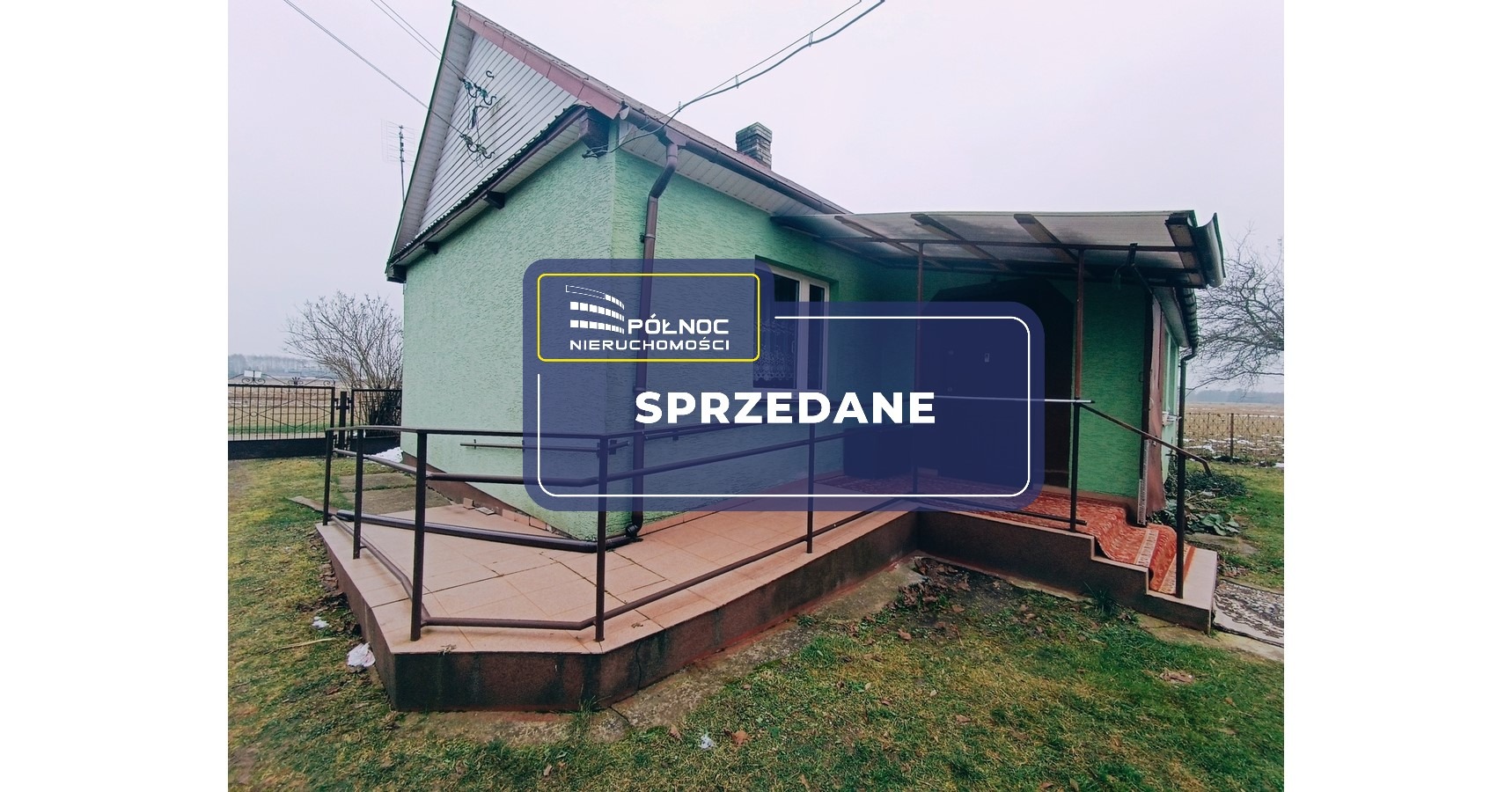 Zdjęcie małego, zielonego domu z werandą, na którym nałożony jest transparent z logo agencji nieruchomości i napisem 'Sprzedane'.