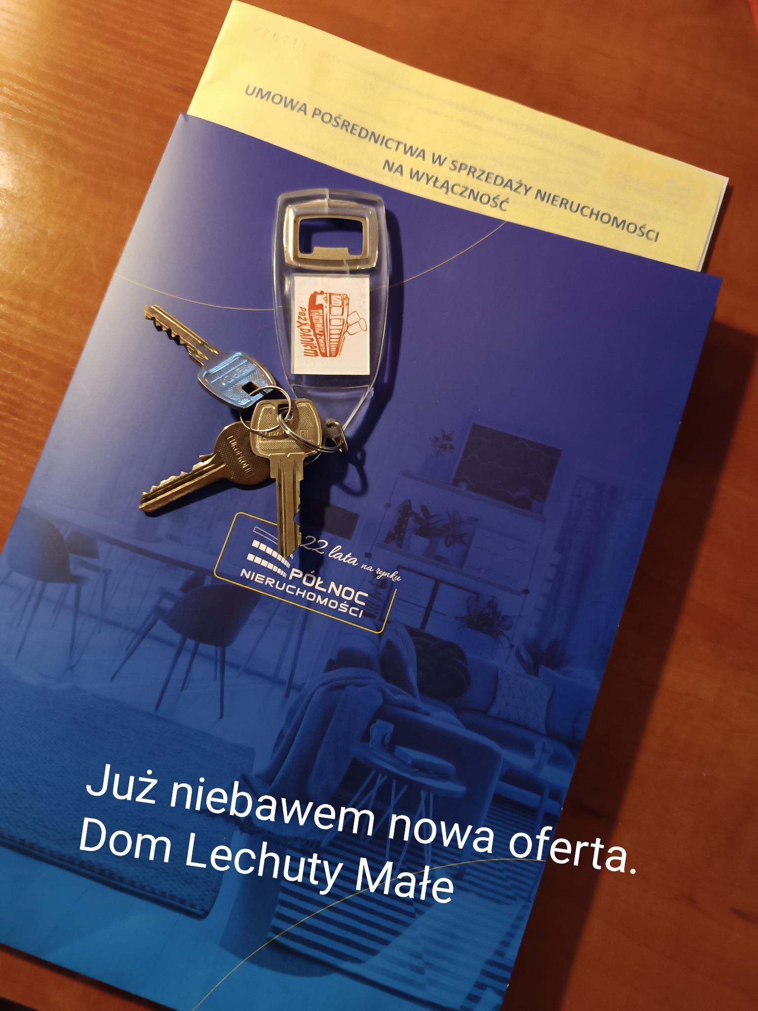 Klucze z brelokiem agencji nieruchomości leżące na folderze reklamowym 'Północ Nieruchomości' i umowie pośrednictwa w sprzedaży nieruchomości na wyłączność.