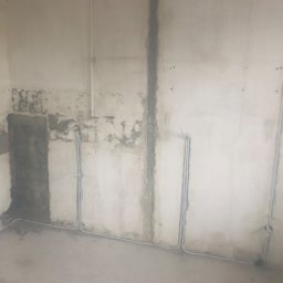 RRS life - Surowa ściana w trakcie remontu z widocznymi kablami elektrycznymi w peszlach, częściowo ukrytymi w bruzdach, z fragmentami starej farby i tynku oraz widoczną rurą instalacyjną pod sufitem.