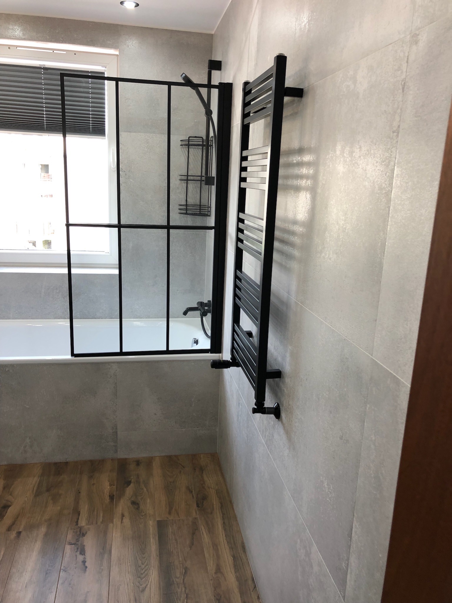 Nowoczesna łazienka z kabiną prysznicową typu walk-in z czarnymi profilami, grzejnikiem drabinkowym w kolorze czarnym i szarymi płytkami imitującymi beton na ścianach, uzupełniona drewnopodobną...