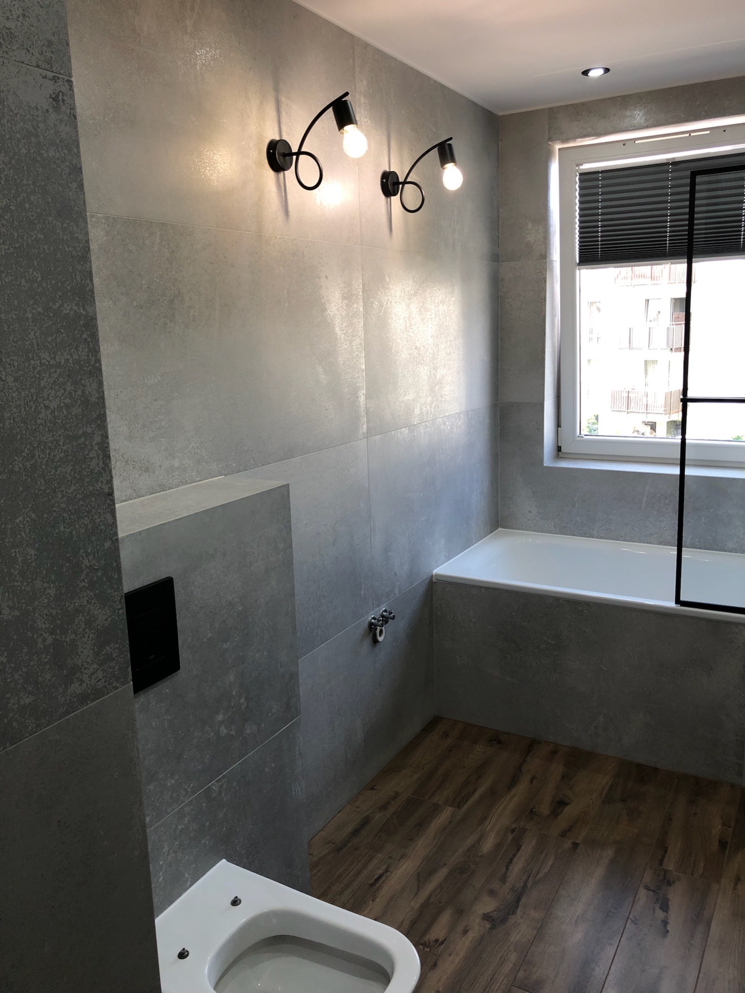 Nowoczesna łazienka z szarymi, matowymi płytkami imitującymi beton, drewnopodobną podłogą, minimalistyczną armaturą i czarnymi lampami ściennymi w stylu industrialnym.