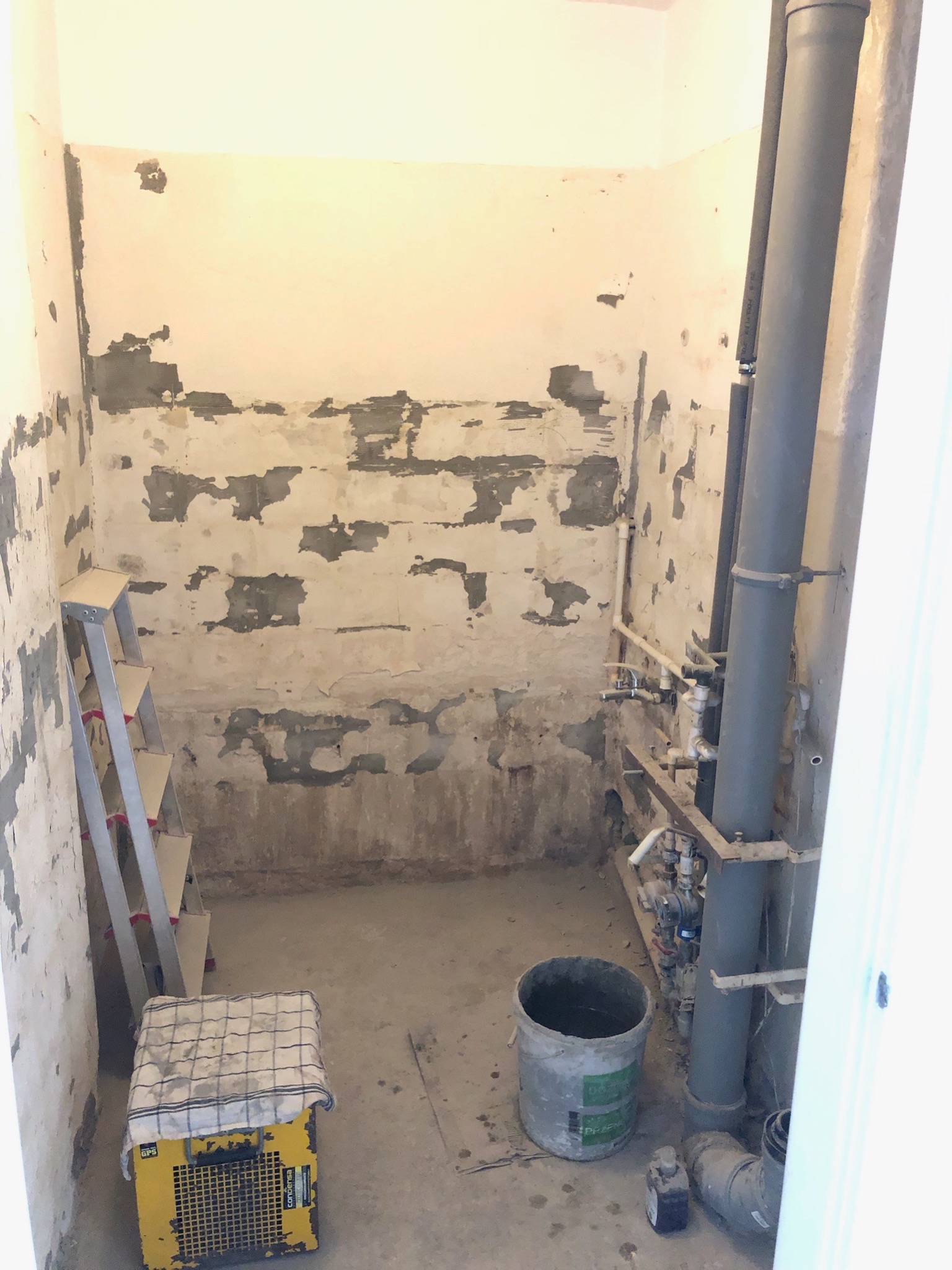 Łazienka w trakcie przygotowania do prac wykończeniowych: otynkowane ściany z widocznymi śladami po starych płytkach, drabina oparta o ścianę, rury instalacyjne, żółty osuszacz budowlany przykryty...