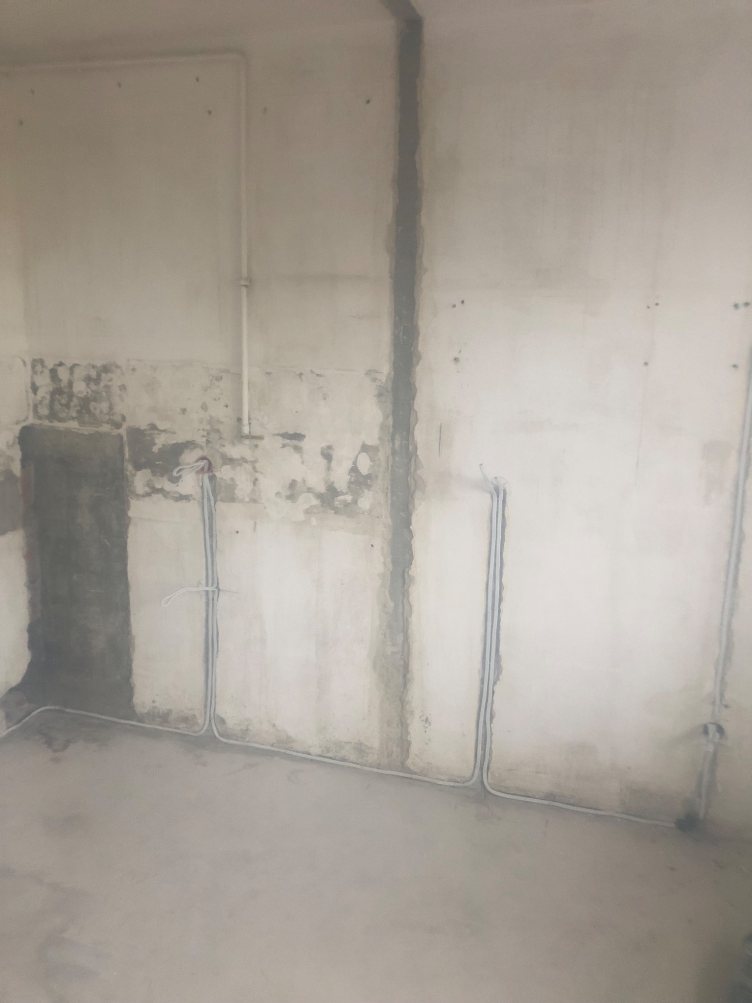 Surowa ściana w trakcie remontu z widocznymi kablami elektrycznymi w peszlach, częściowo ukrytymi w bruzdach, z fragmentami starej farby i tynku oraz widoczną rurą instalacyjną pod sufitem.