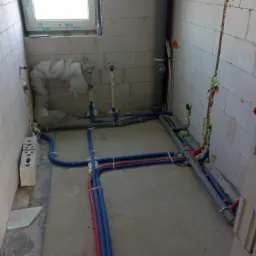 Surowy stan łazienki z widoczną instalacją wodną i kanalizacyjną w trakcie montażu, rury prowadzone po podłodze i w ścianach z bloczków, zabezpieczone pianką montażową.