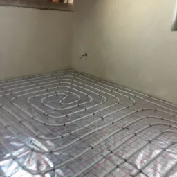 Rozłożona instalacja ogrzewania podłogowego z rurkami na folii izolacyjnej z czerwoną siatką, widoczne elementy mocujące, surowa ściana i fragment drewnianej konstrukcji stropu.