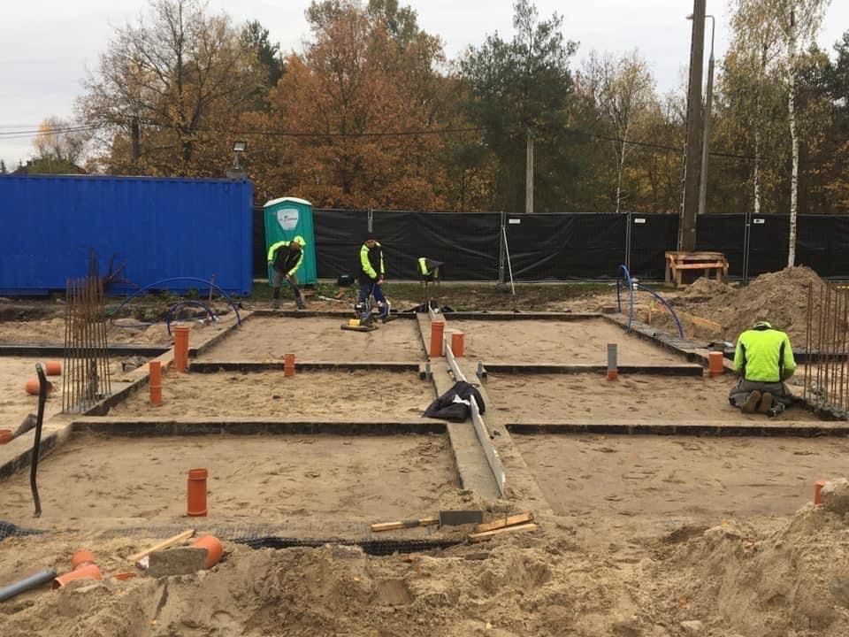 Przygotowanie fundamentów pod budynek: pracownicy w odblaskowych ubraniach układają rury kanalizacyjne w wykopie, widoczne szalunki i zbrojenia, na drugim planie kontener budowlany i przenośna...