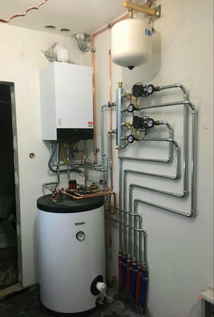 Nowoczesny kocioł grzewczy z zasobnikiem ciepłej wody i rozbudowaną instalacją hydrauliczną z rur miedzianych i stalowych, manometry na pionowej rurze, widok z boku, surowy stan wykończenia ścian.
