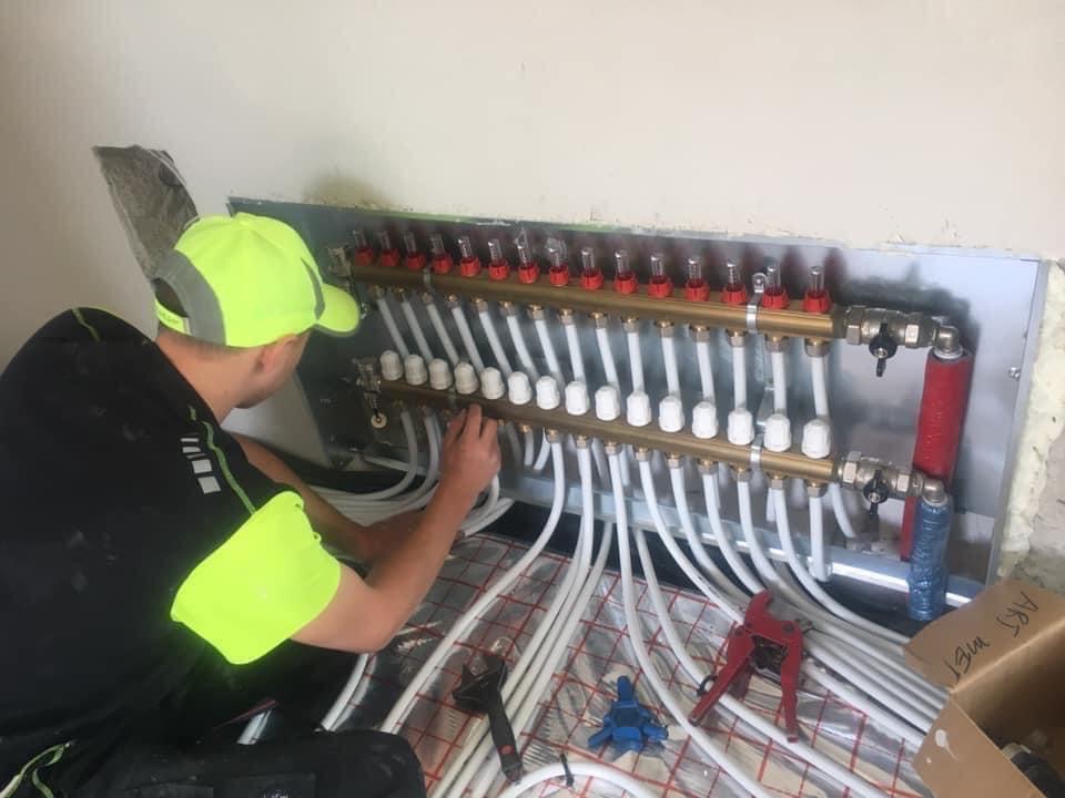 Instalator w neonowej kamizelce podłącza rury do rozdzielacza ogrzewania podłogowego, widoczne narzędzia i fragment izolacji podłogowej.