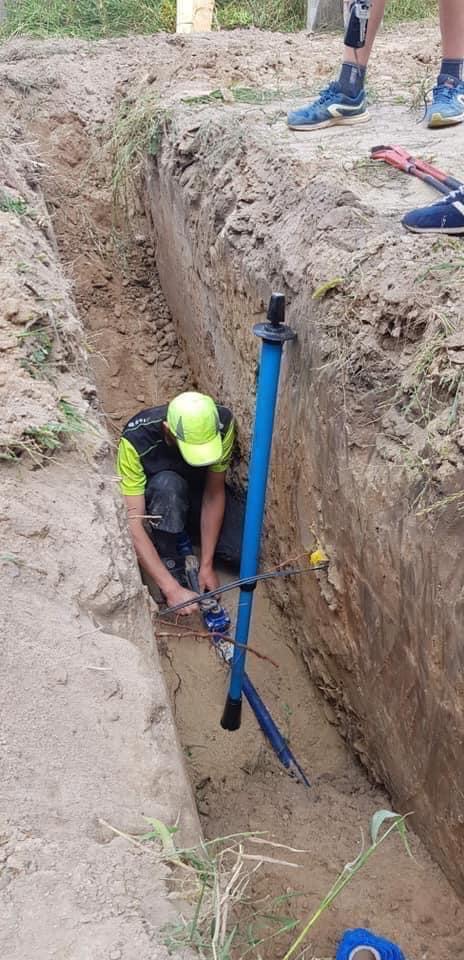 Pracownik w wykopie łączy elementy instalacji wodociągowej, obok niebieska rura i narzędzia, widoczne ślady ziemi na ubraniu.