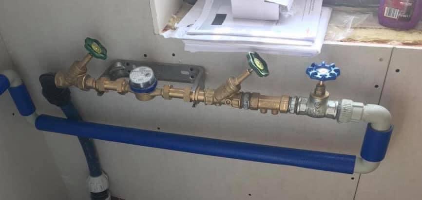 Zestaw rur wodociągowych z zaworami (zielonymi i niebieskim) oraz licznikiem wody, zamontowany na ścianie wykończonej płytą gipsowo-kartonową. Widoczne połączenia mosiężne i elementy z tworzywa...