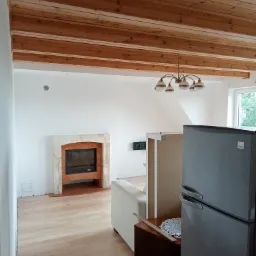 Wnętrze pokoju z drewnianym sufitem, jasnymi ścianami i marmurowym kominkiem, widoczne fragmenty mebli i sprzętu AGD, podłoga wyłożona panelami.