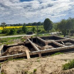KD EXPERT Konrad Drzazga - Fundamenty domu w trakcie budowy, widoczne szalunki i izolacja, w tle krajobraz z polem rzepaku i przejeżdżający samochód.