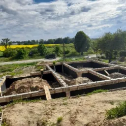 Fundamenty domu w trakcie budowy, widoczne szalunki i izolacja, w tle krajobraz z polem rzepaku i przejeżdżający samochód.